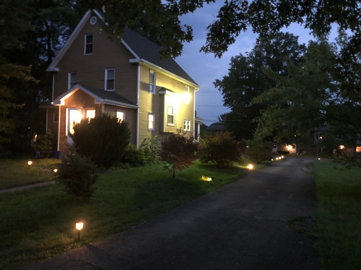 The turn-of-the-century farmhouse is softly illuminated at dusk, showcasing a welcoming façade with light streaming from the front windows. A well-maintained lawn features garden plants, and pathway lights guide the way along the driveway, leading to the entrance.