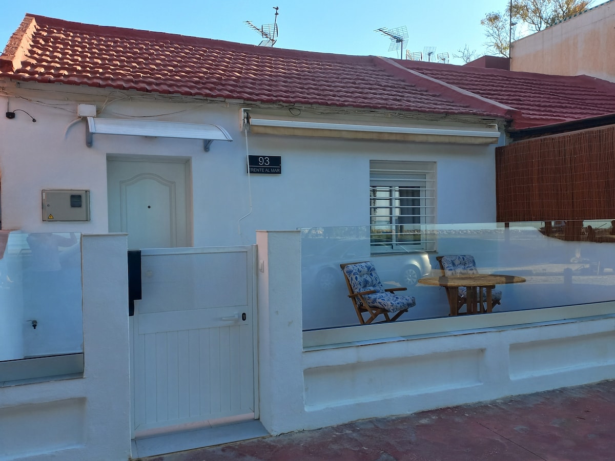 A single-story white house features a welcoming entrance with a glass barrier at the front. Two wooden chairs are arranged beside a small table, providing a cozy outdoor seating area. The roof is tiled, and details like a house number plaque are visible.