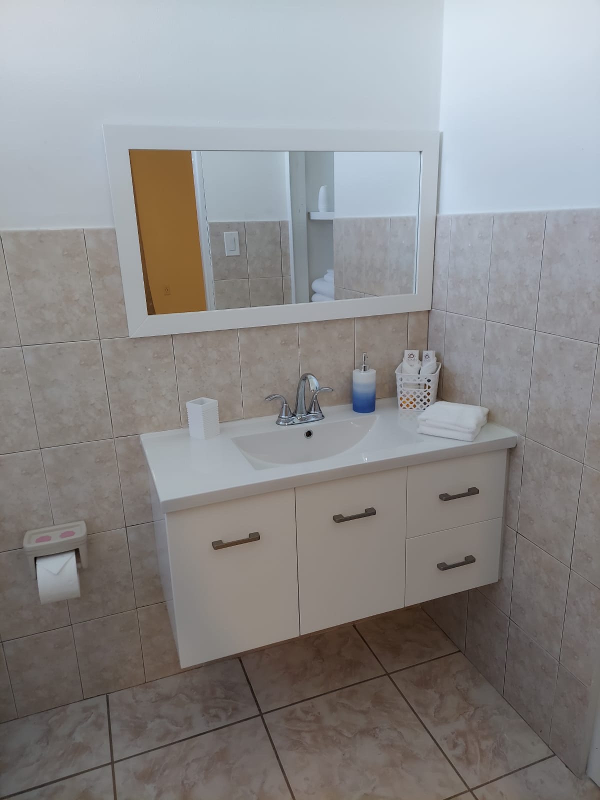 A clean and well-lit bathroom features a compact vanity with a sink and modern fixtures. Soft towels are neatly arranged beside a soap dispenser. A mirror above the sink reflects the tiled walls, while a toilet tissue holder is positioned nearby for convenience.