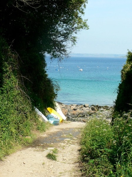 A narrow pathway, bordered by lush greenery and trees, leads down to a tranquil beach scene. Brightly colored kayaks rest on the sandy shore, while calm turquoise waters stretch out, meeting a clear blue sky. Small boats can be seen in the distance.