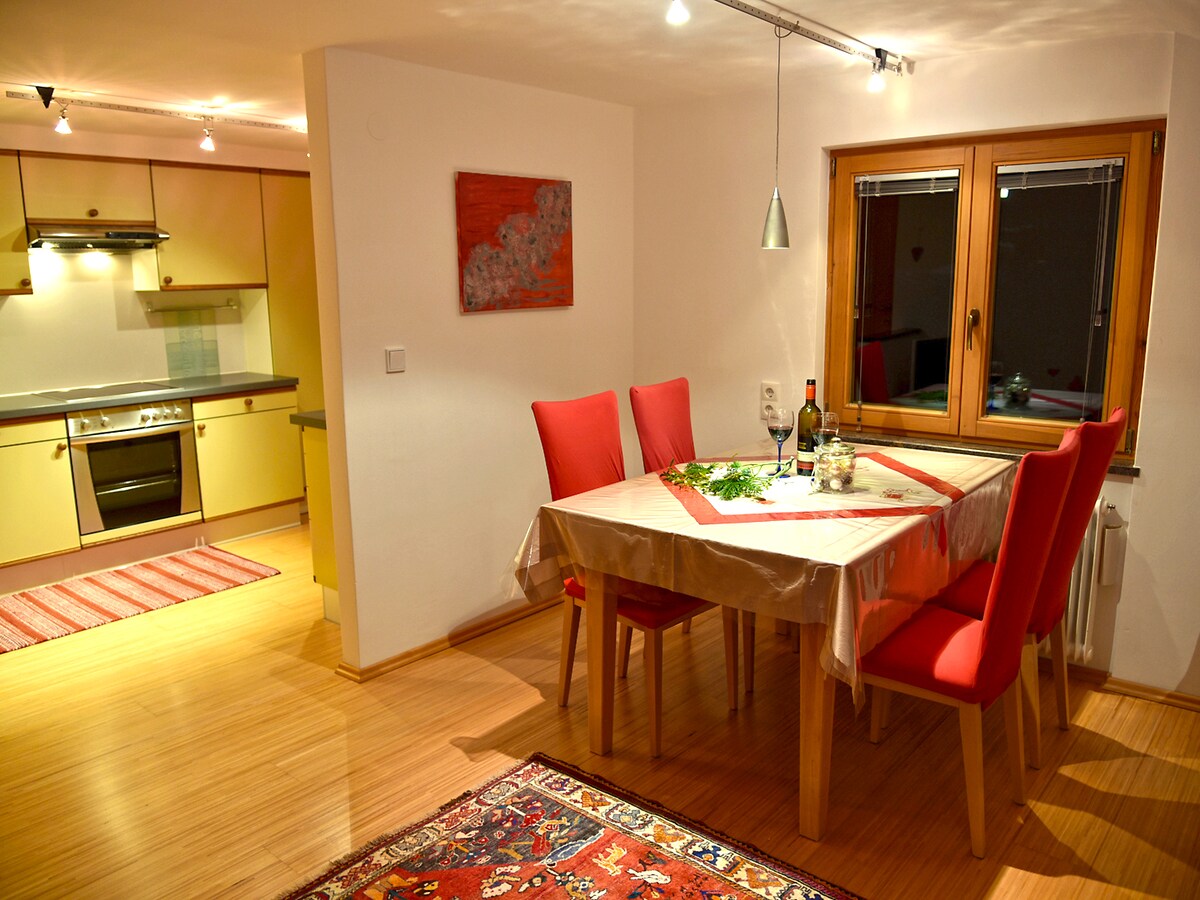 A dining area features a wooden table surrounded by red chairs. Glassware and a bottle are placed on the table, complemented by a decorative centerpiece. A kitchen is visible in the background, showcasing modern appliances and cabinetry. Natural light enters through a window dressed with a simple covering.