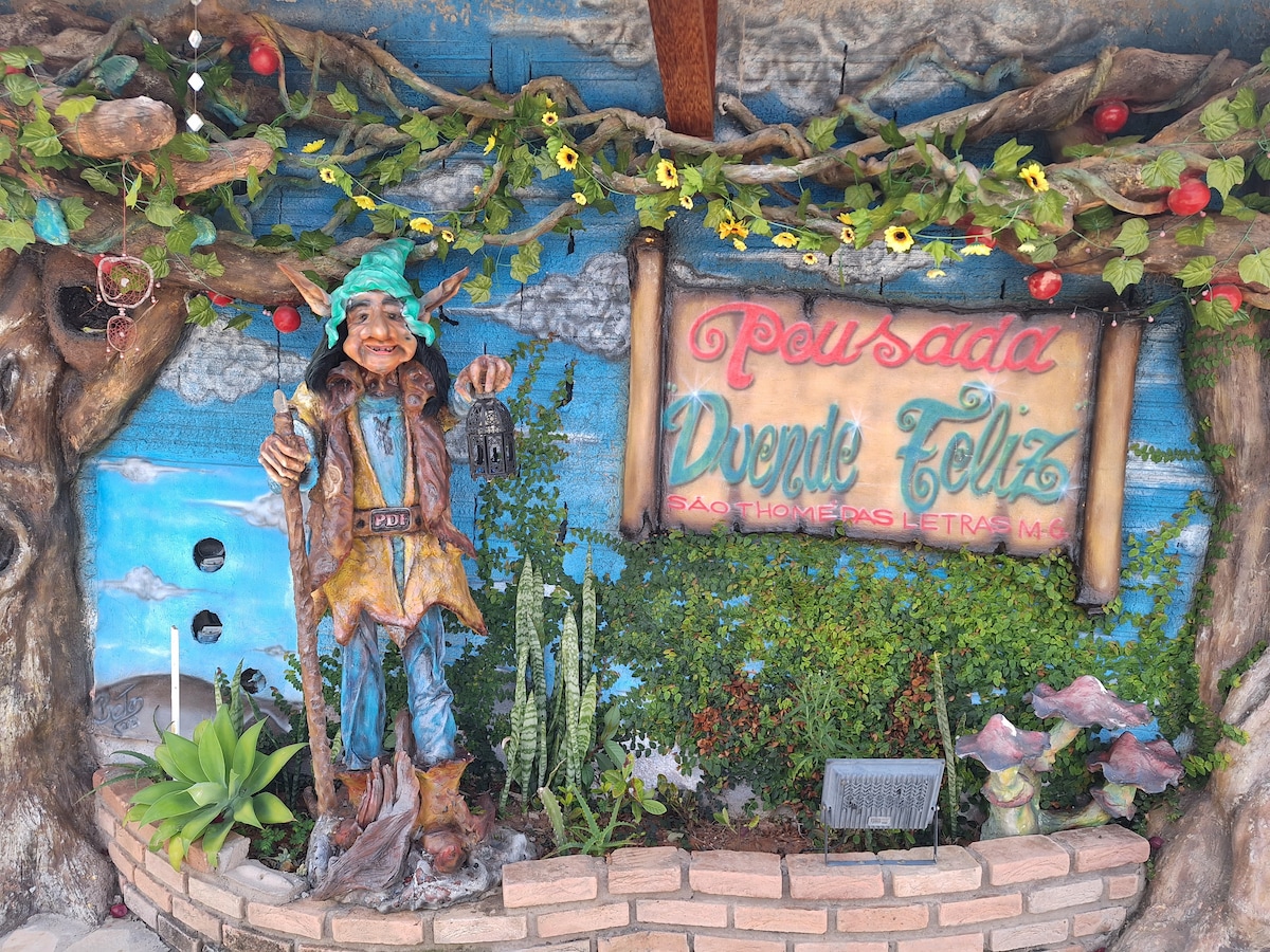 An outdoor entryway features a colorful mural depicting a serene landscape. A wooden sign is prominently displayed, identifying the property. A decorative statue of a figure holding a lantern stands beside it, surrounded by various plants and flowers that add to the welcoming atmosphere.