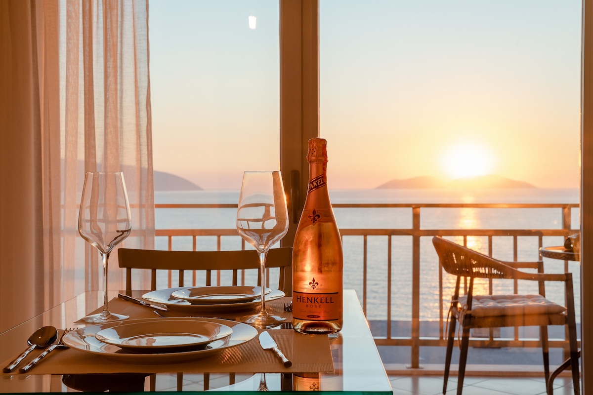 A dining table is set with elegant glassware and plates, accompanied by a bottle of Henkell sparkling wine. Soft sunlight filters through sheer curtains, illuminating the space, while a scenic view of the sea and sunset can be seen through large glass doors.