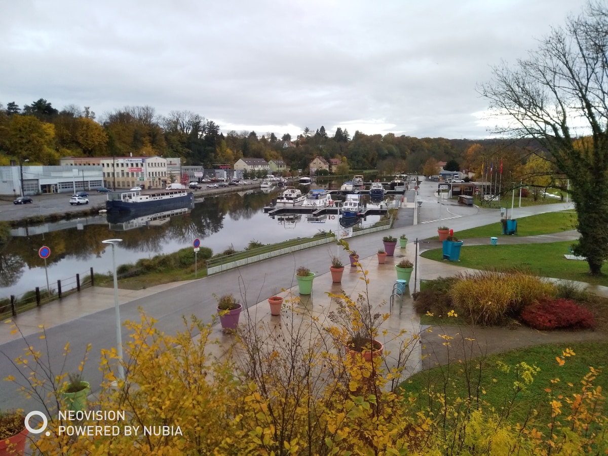 A scenic view of a calm port area with various boats moored along the water. The waterfront is lined with trees and colorful flower pots. Soft reflections of the buildings and nature are seen in the water, creating a serene atmosphere.