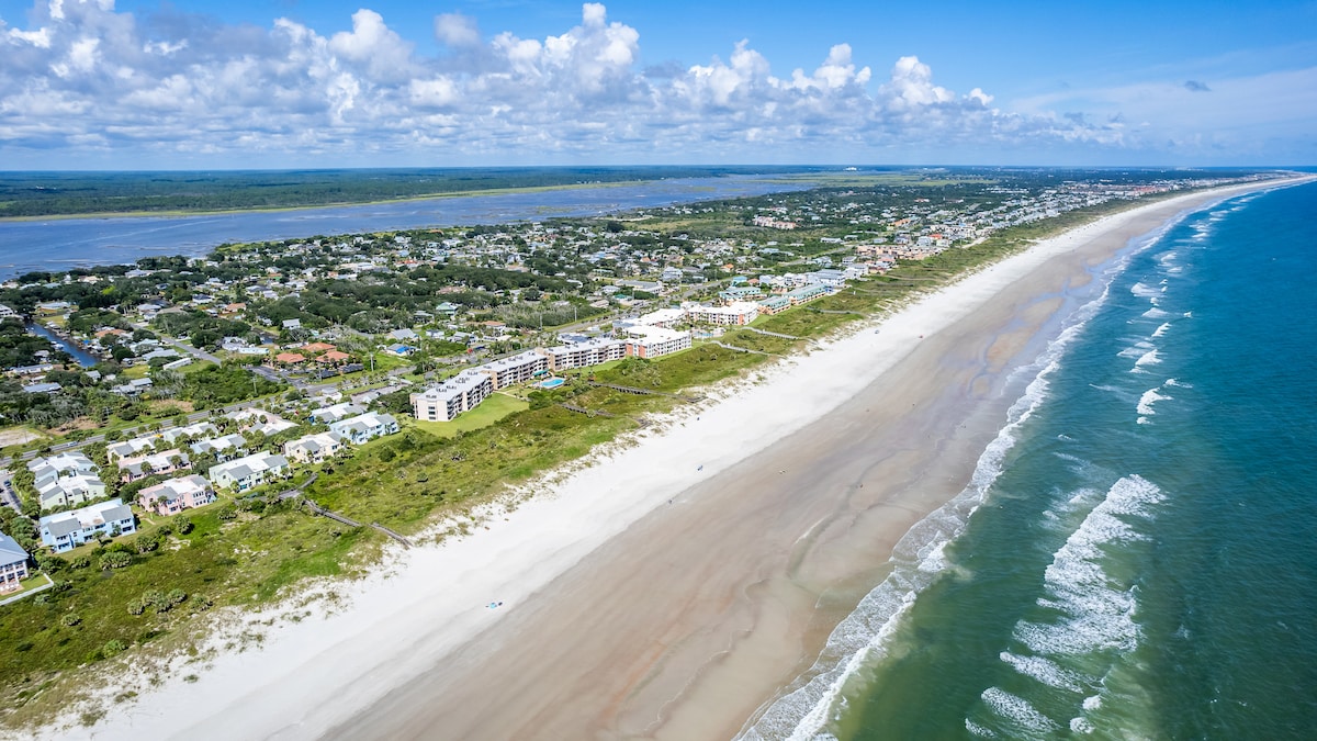An aerial view captures the expansive coastline along with the sandy beach and gentle waves of the ocean. Surrounding areas showcase lush greenery and residential buildings, highlighting the serene landscape under a clear blue sky with scattered clouds.