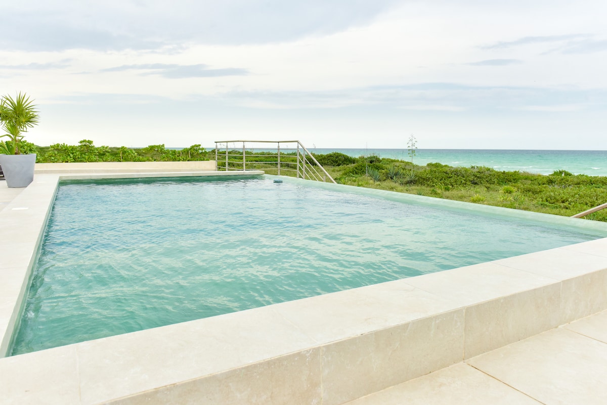 An outdoor swimming pool with clear water reflects the surrounding greenery and ocean views. A stainless steel railing provides accessibility to the pool area, while gentle waves can be seen in the distance against a cloudy sky.