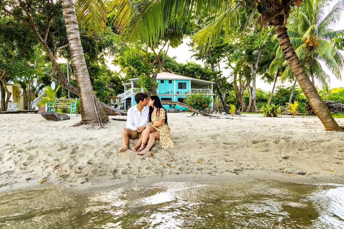 A couple is seated on the sandy beach, framed by palm trees, enjoying a moment together. A colorful building is seen in the background, surrounded by lush greenery. The water gently laps at the shore, creating a serene coastal scene.