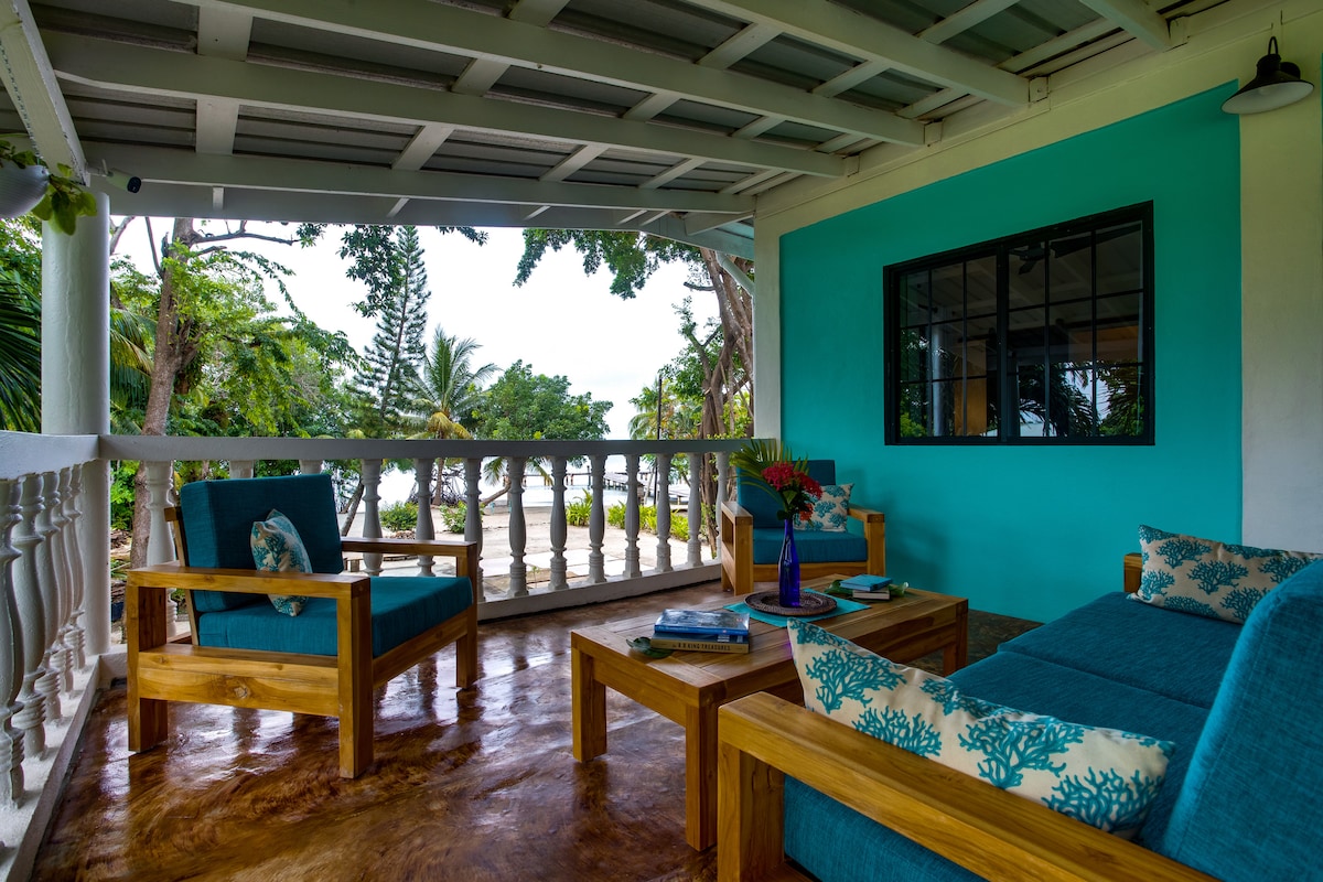 A spacious verandah is furnished with wooden seating and tables, featuring light blue cushions that complement the surrounding tropical landscape. Potted flowers and coastal decor add detail, while views of the lush greenery and distant ocean create a serene atmosphere.