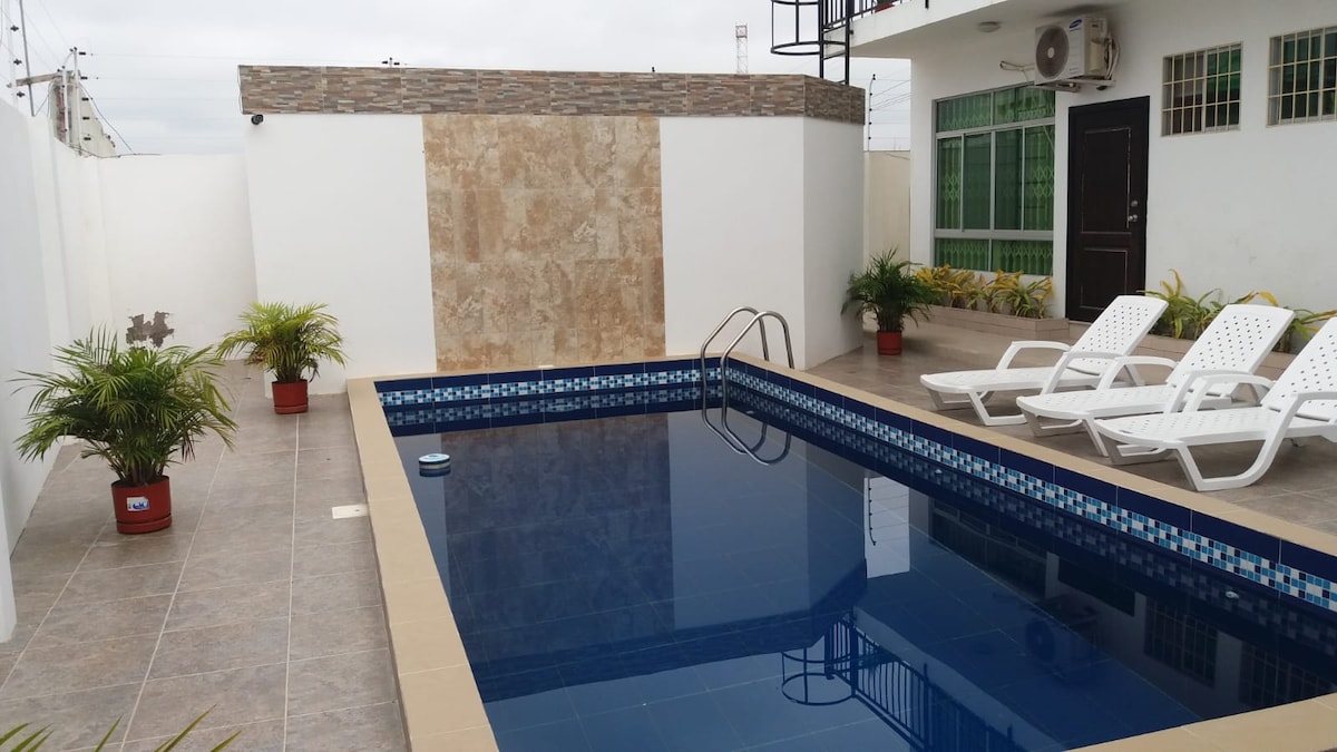 A well-maintained swimming pool is showcased, surrounded by a tiled patio. Two white lounge chairs are positioned nearby, and potted plants add greenery to the space. The pool features a blue mosaic border and is complemented by a textured stone wall in the background.