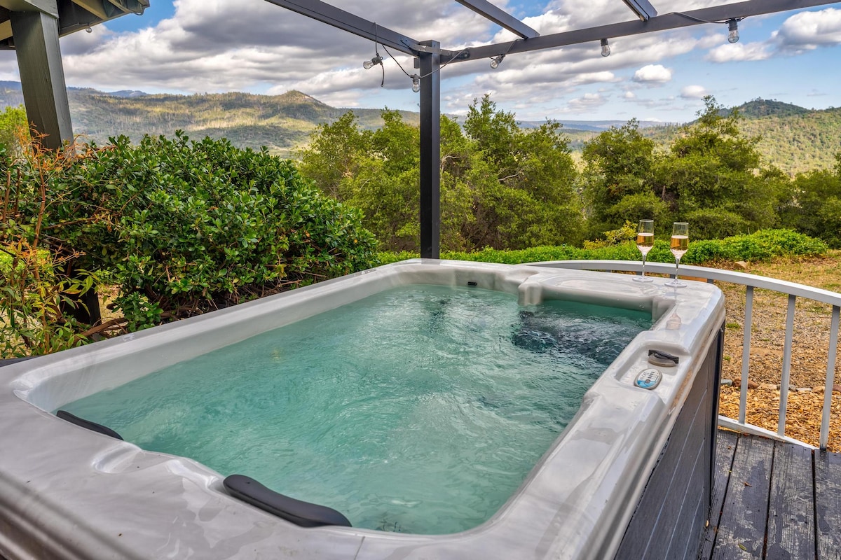A hot tub is situated on a deck, surrounded by greenery and offering a view of the Sierra Mountain range. Two glasses are placed on the edge of the tub, reflecting a serene outdoor setting under a partly cloudy sky.