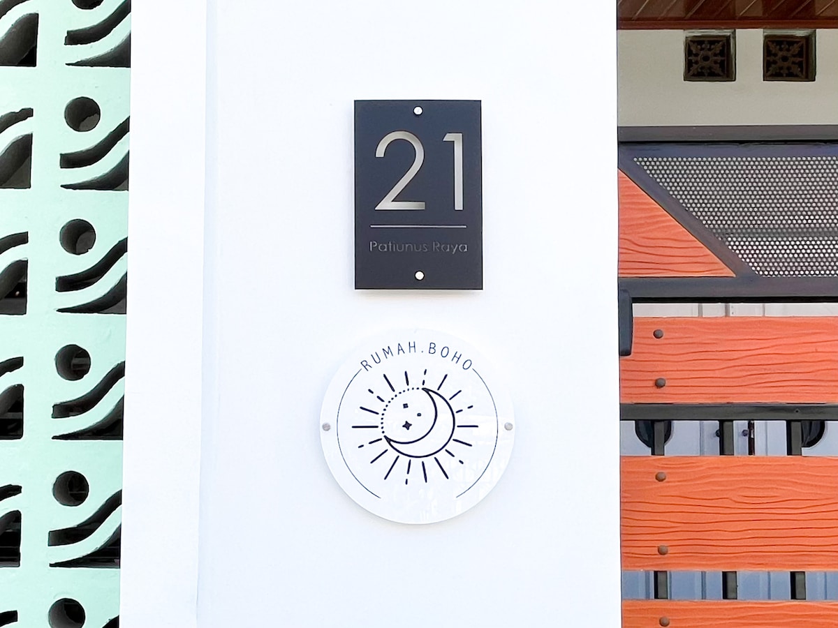 A clear view of the property number '21' displayed on an elegant black sign. Below, a round sign features the logo 'RUMAH BOHO' with a sun design, positioned against a white wall. The entryway design includes decorative elements enhancing its distinctive style.