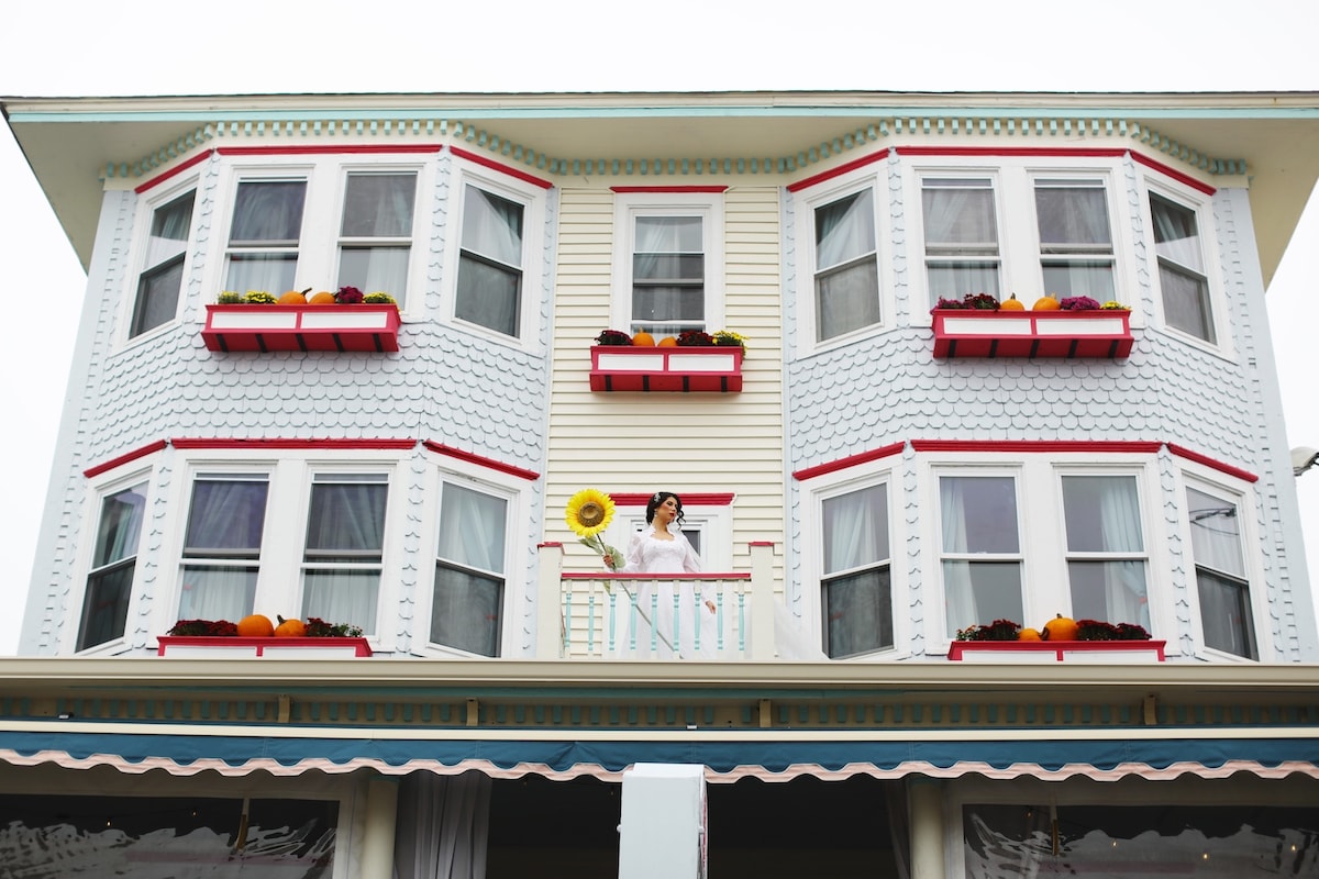 The exterior of a Victorian-style building features three large bay windows adorned with colorful flower boxes. A person stands on the balcony holding a sunflower, while intricate architectural details highlight the charm of the structure.