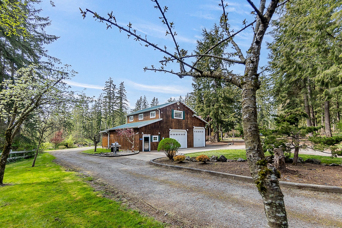 A modern barn home is set amidst a lush forest, featuring a gravel driveway that leads to its front. Large garage doors can be seen, along with landscaped areas marked by vibrant greenery and scattered foliage.