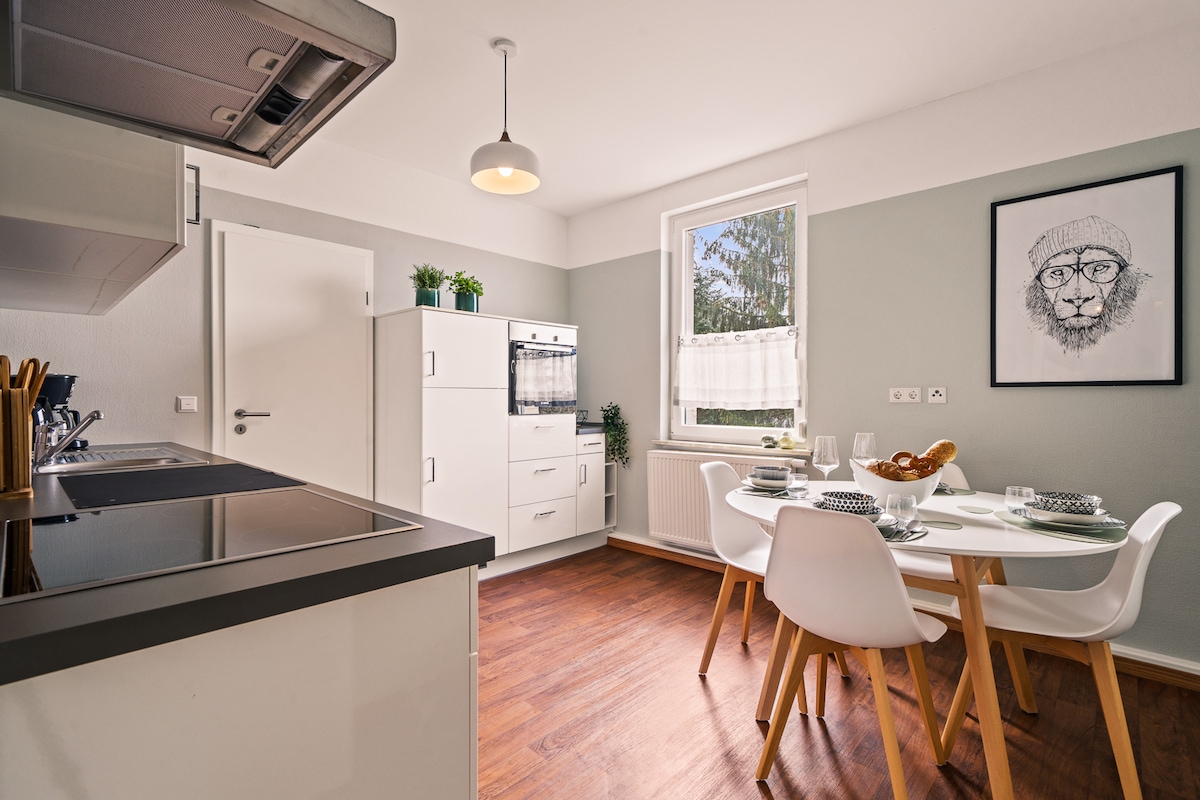 An inviting kitchen is showcased, featuring modern appliances including an oven and stovetop. A dining area with a round table is surrounded by four white chairs. Natural light enters through a window, complementing the subtle green and white color scheme.