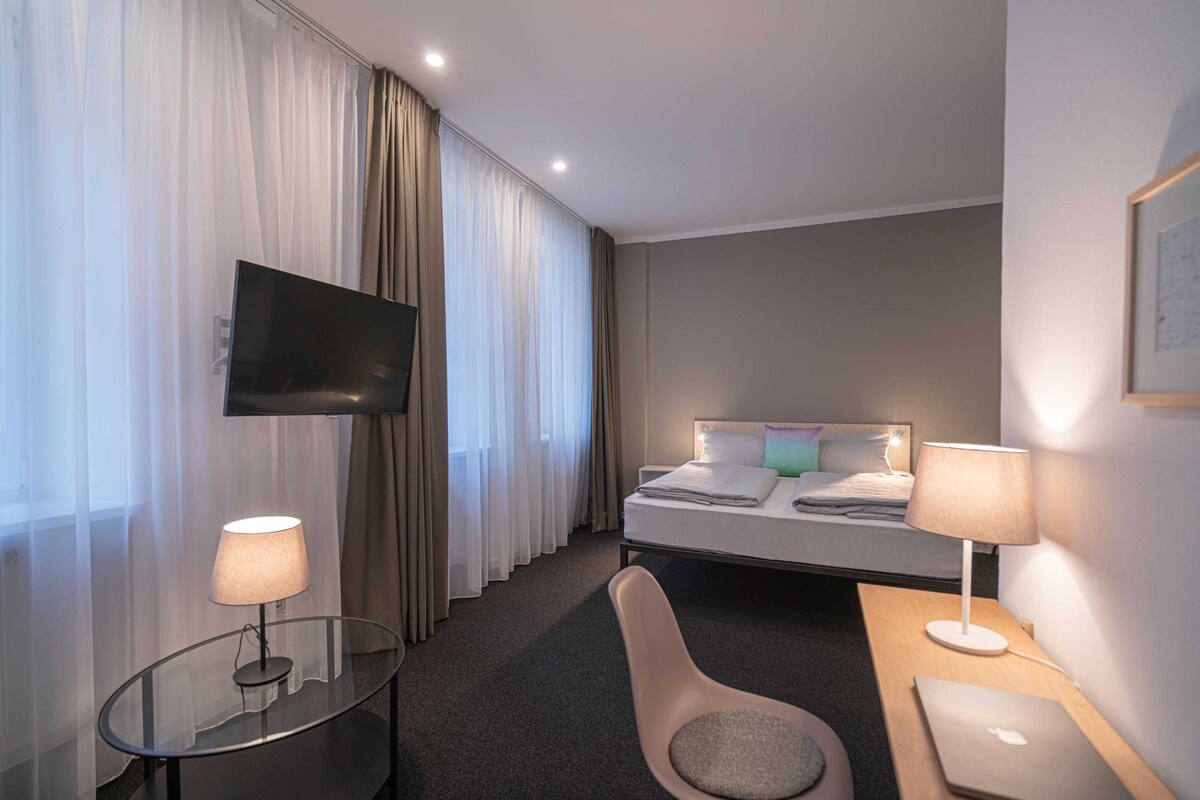 A spacious hotel room features large windows adorned with sheer curtains, allowing natural light to filter in. A modern bed is positioned against a neutral wall, accompanied by a TV mounted on the opposite side. A small coffee table and desk complete the layout, providing functional spaces.