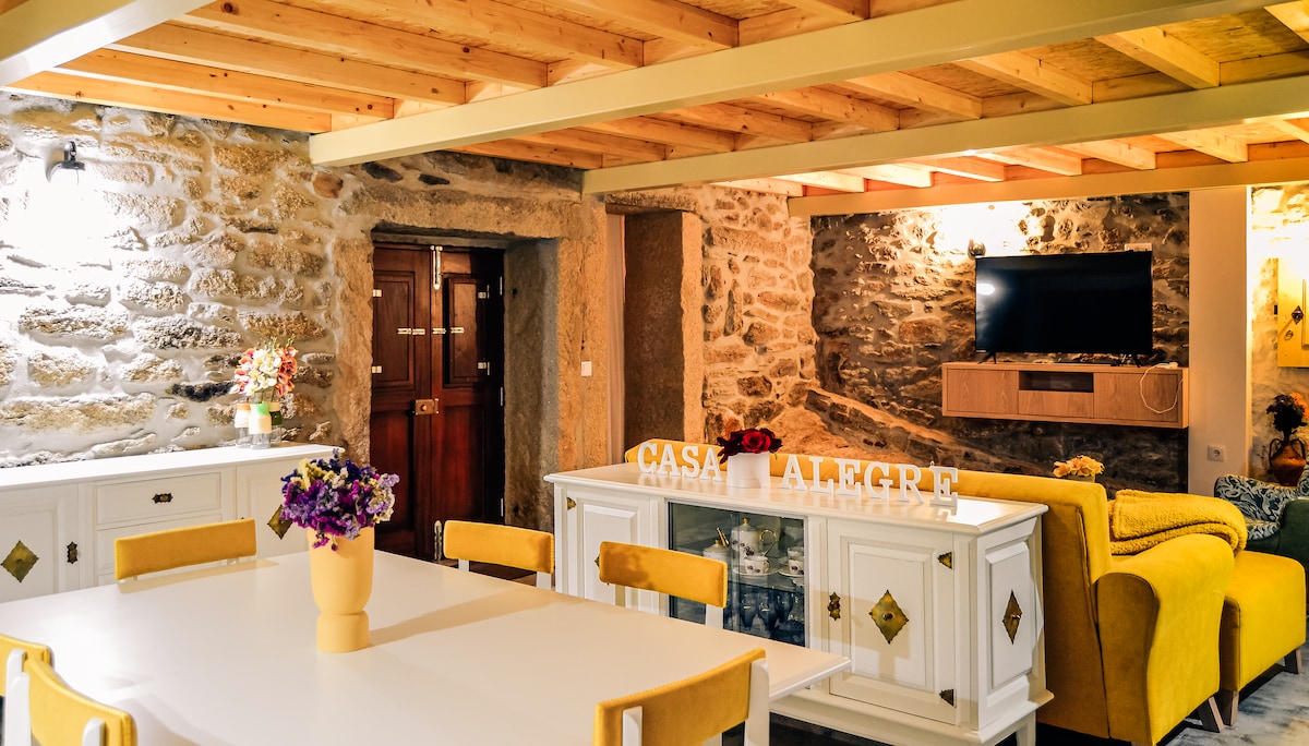 A welcoming living and dining area features rustic stone walls and wooden beams. A large white dining table is centered beneath warm lighting, surrounded by stylish chairs. A cozy yellow sofa is positioned nearby, facing a wall-mounted television, creating a relaxed environment for gatherings.