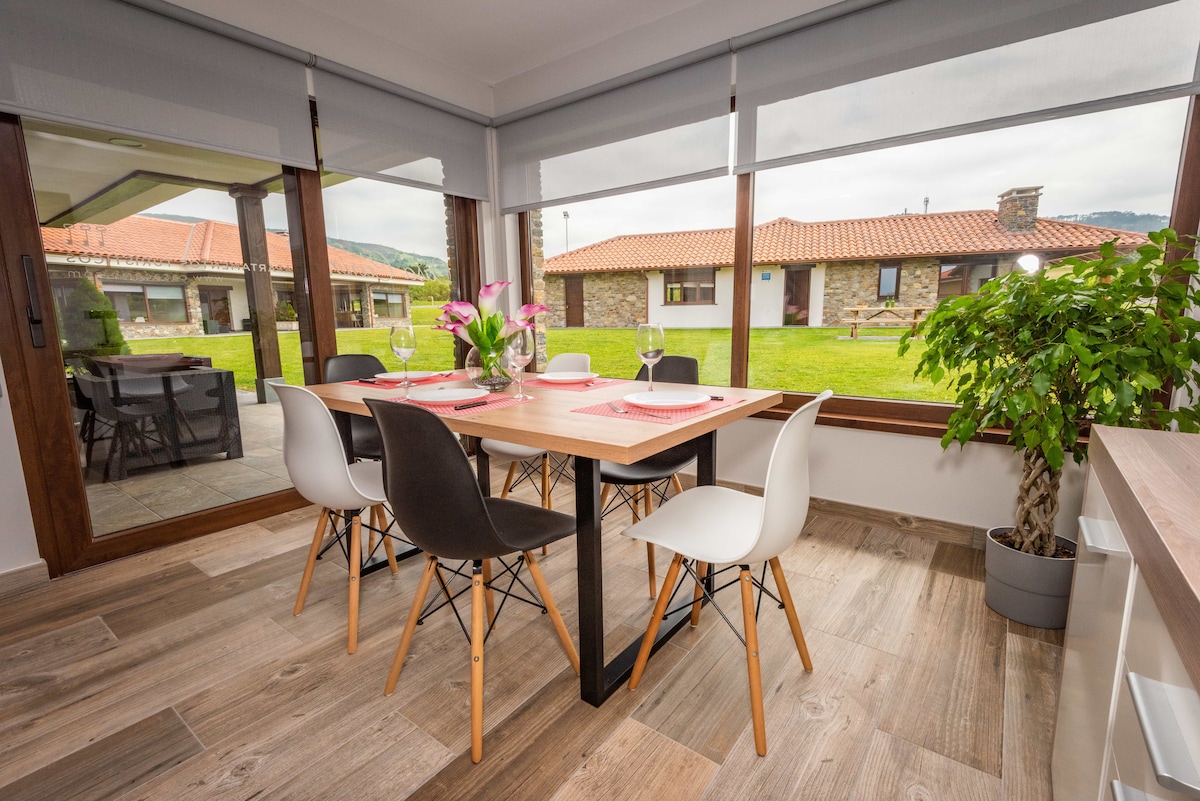 A dining area features a modern table set for six, complemented by a variety of stylish chairs. Large windows invite natural light and offer views of a green outdoor space and stone buildings with terracotta roofs.