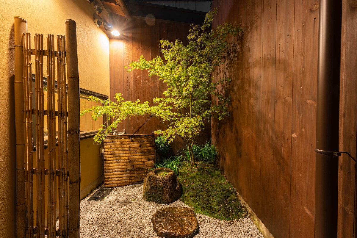 A serene courtyard features a small stone basin surrounded by carefully arranged gravel. A vibrant green tree stands in the corner, offering a touch of nature, while wooden elements create a calm and harmonious atmosphere.