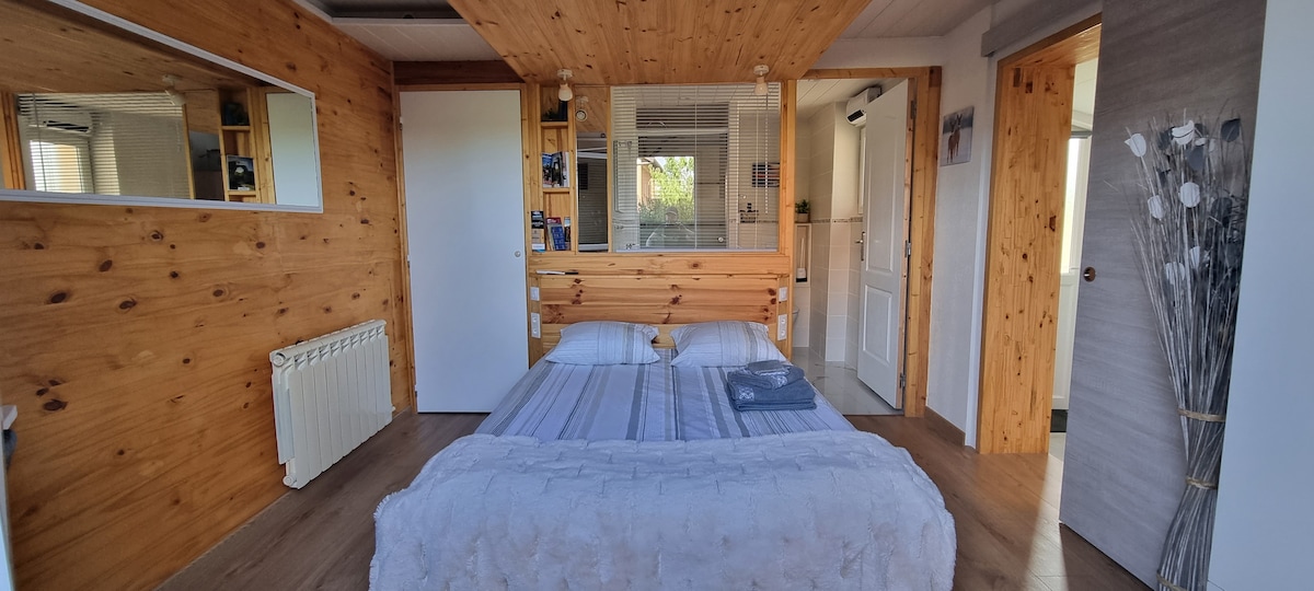 A cozy sleeping area features warm wooden walls and soft lighting. A spacious bed with light linens is set against a wooden headboard. Shelving units and a mirror are visible, while a decorative element of twigs adds a touch of nature to the room.