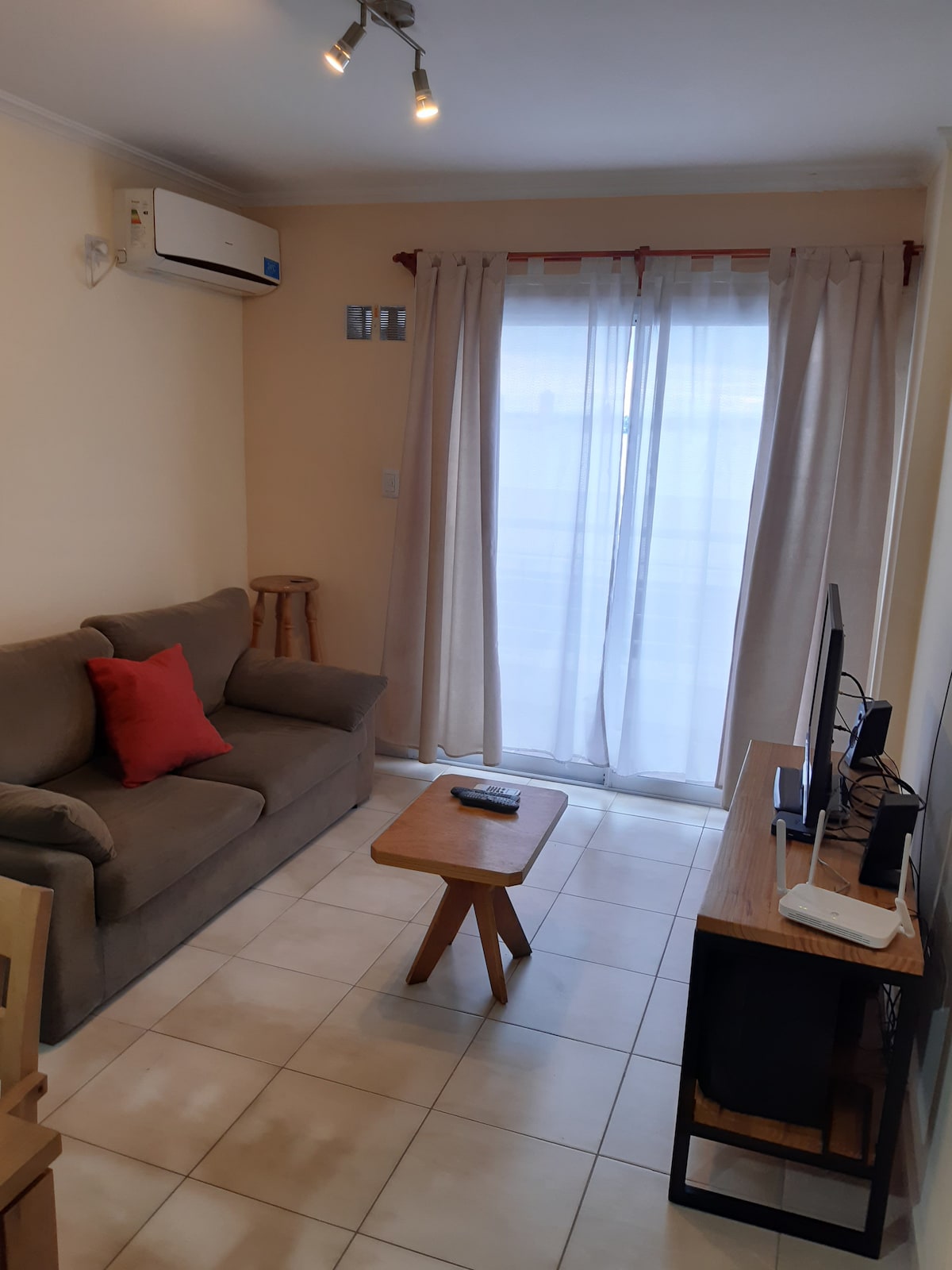 The living area is furnished with a comfortable sofa and a small wooden coffee table. Natural light fills the space through a large window with sheer curtains, providing a bright ambiance. An air conditioning unit is mounted on the wall for climate control.