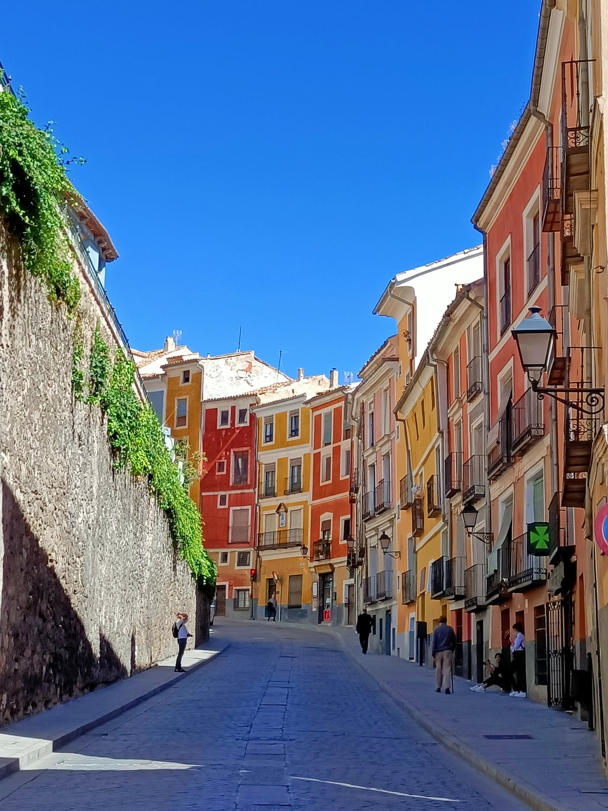 A charming street in the historical district is lined with buildings showcasing various warm tones, creating a vibrant atmosphere. Green foliage drapes over a stone wall, while a gentle incline leads towards the blue sky. Pedestrians stroll along the cobblestone pathway, embodying the area's lively spirit.