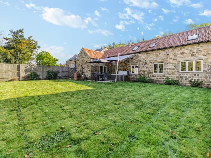 A spacious garden area is shown, featuring well-maintained grass with clear mowing lines. The stone exterior of the property is visible, with multiple windows allowing natural light. A sheltered outdoor seating area and dining table are situated on the patio, surrounded by greenery.