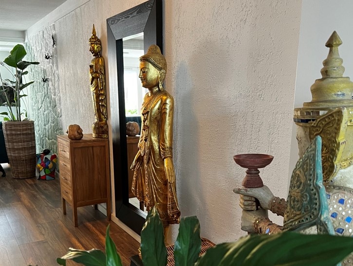 A serene interior space is highlighted by golden statues, including a standing figure and a seated Buddha, creating a calming ambiance. A tall mirror reflects the decor, while lush green plants and an accent piece contribute to the tranquil atmosphere.