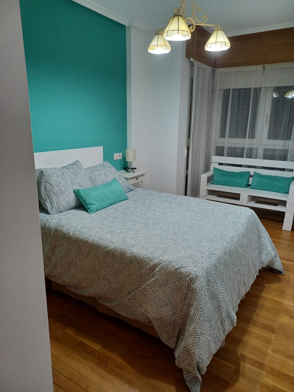 The bedroom features a spacious bed with a patterned duvet and two accent cushions. A cozy seating area is positioned next to the window, adorned with turquoise cushions. Soft lighting is provided by a three-bulb chandelier overhead, enhancing the calming color scheme.