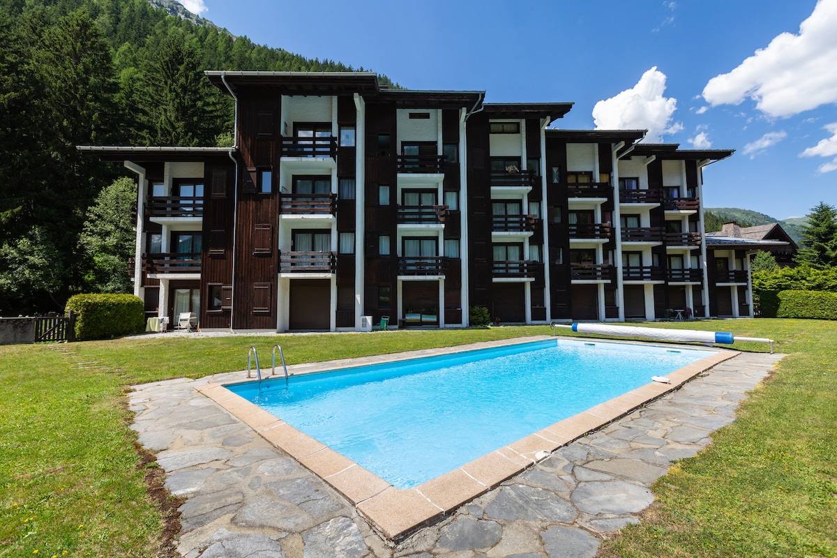 A modern condominium building with multiple balconies overlooks a well-maintained garden area. In the foreground, a clear swimming pool is framed by a stone patio, inviting relaxation in the summertime. Lush greenery and mountains are visible in the background.