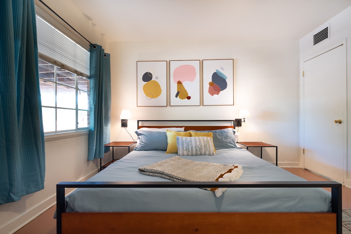 A cozy bedroom is highlighted by a spacious bed with soft blue linens and decorative pillows. Original artwork is displayed above the headboard, and natural light filters through a window with turquoise curtains. Nightstands are positioned on either side, each with a lamp.