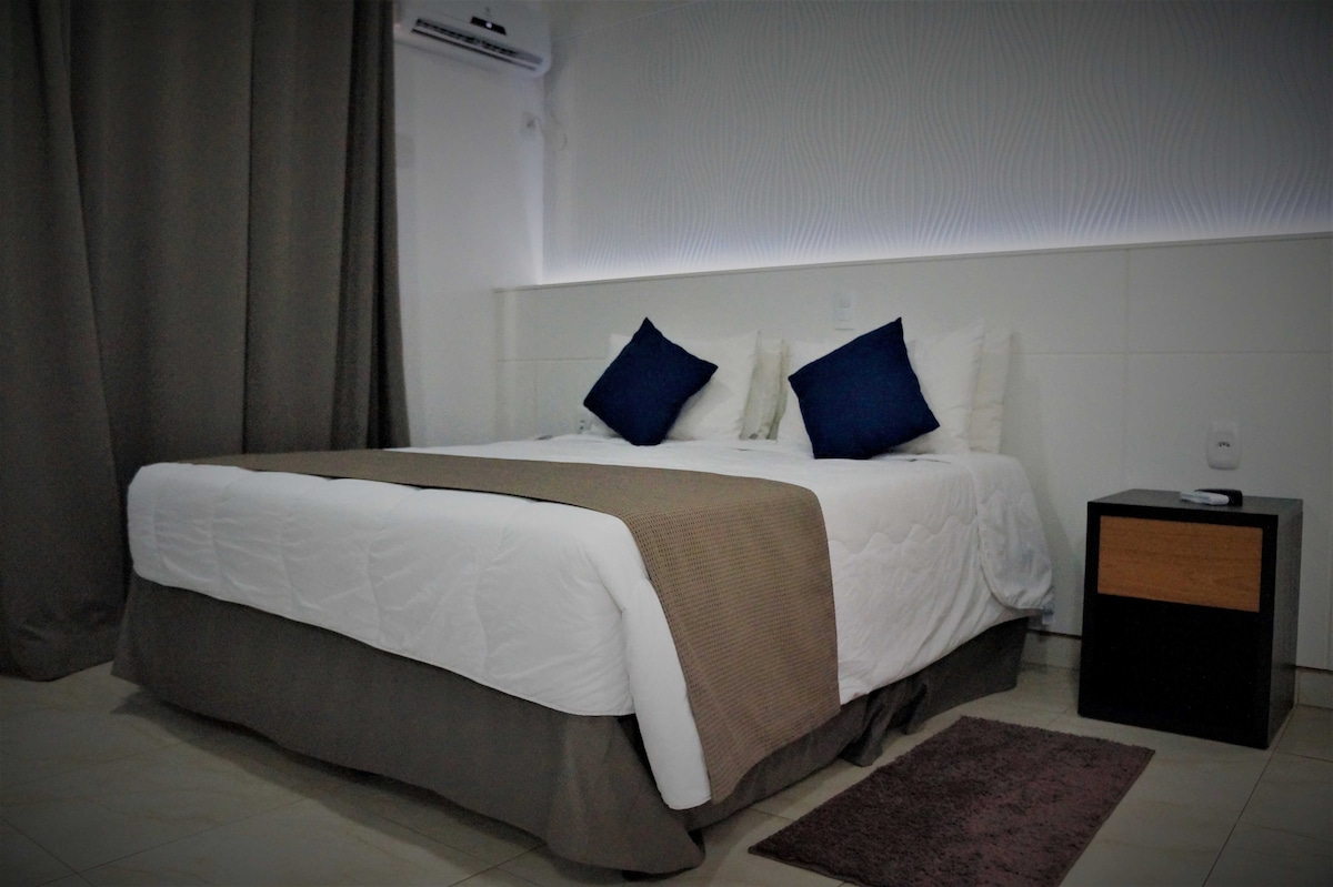 A spacious bed is flanked by two blue accent pillows, set against a softly illuminated headboard. Light-colored bedding contrasts with a dark bedside table, which holds a minimalistic lamp. Neutral curtains frame the window, and a small rug is placed beside the bed.