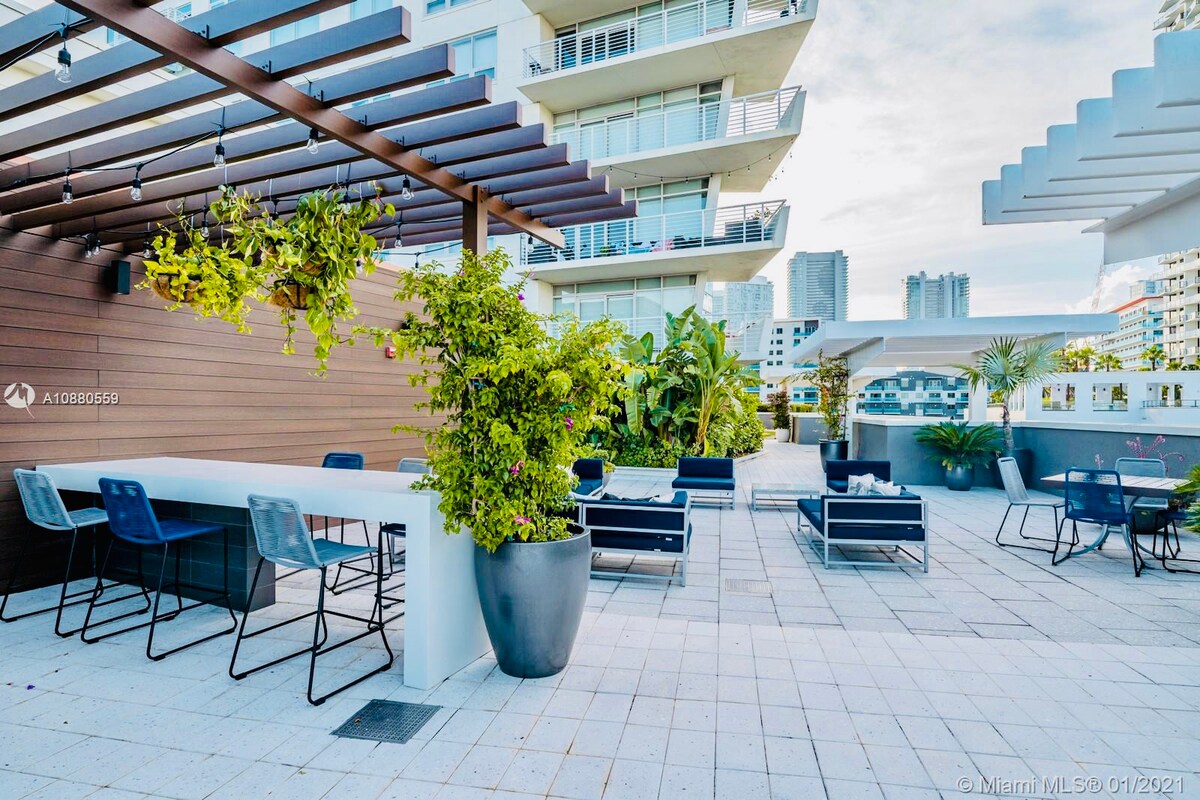 An outdoor terrace features modern seating arrangements and a long countertop for dining. Lush greenery and decorative plants create a refreshing ambiance, with the backdrop showcasing the surrounding high-rise buildings under a bright sky.
