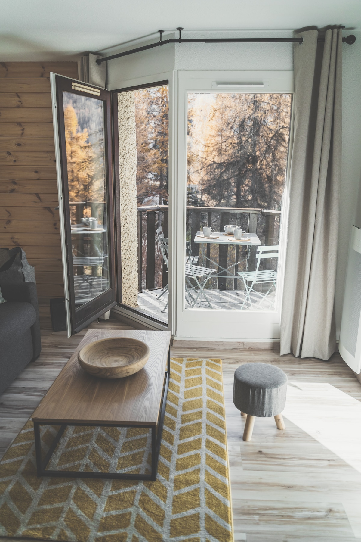 A bright living area features an open door leading to a balcony with outdoor seating. A wooden coffee table rests on a patterned rug, with a bowl placed on top. Natural light illuminates the space, highlighting the warm wood accents.