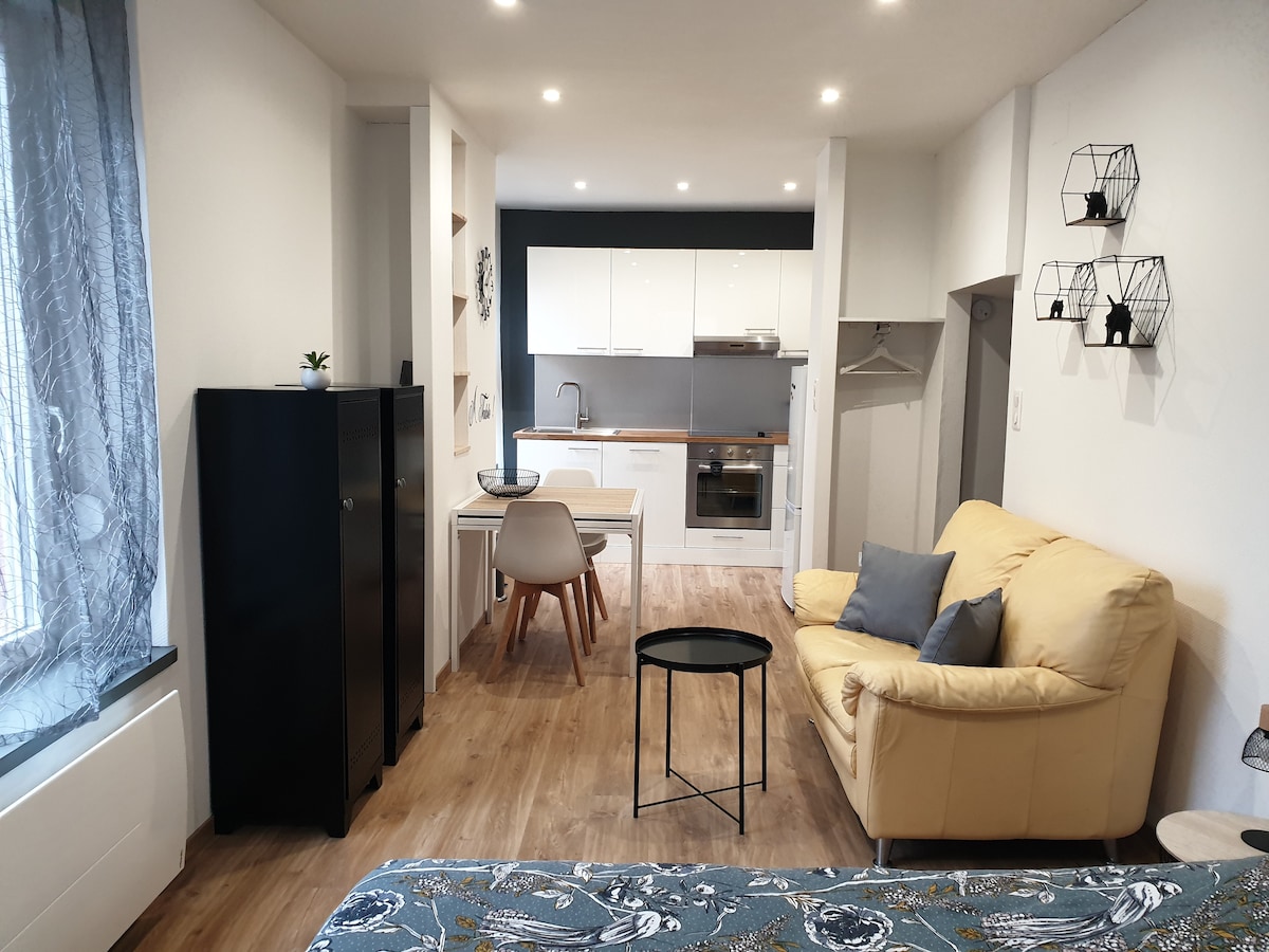 A bright open space features a smoothly designed kitchen with essential appliances, a dining table, and a cozy seating area with a pale yellow sofa. Light wooden flooring complements the decor, while large windows allow ample natural light to brighten the room.