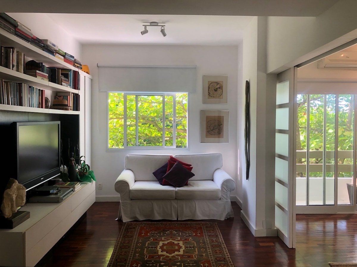 A bright living area features a white sofa adorned with colorful cushions, positioned near a large window that provides views of lush greenery. A low white media console displays decorative items, alongside a cozy area rug on polished wooden flooring. Shelves filled with books line one wall.