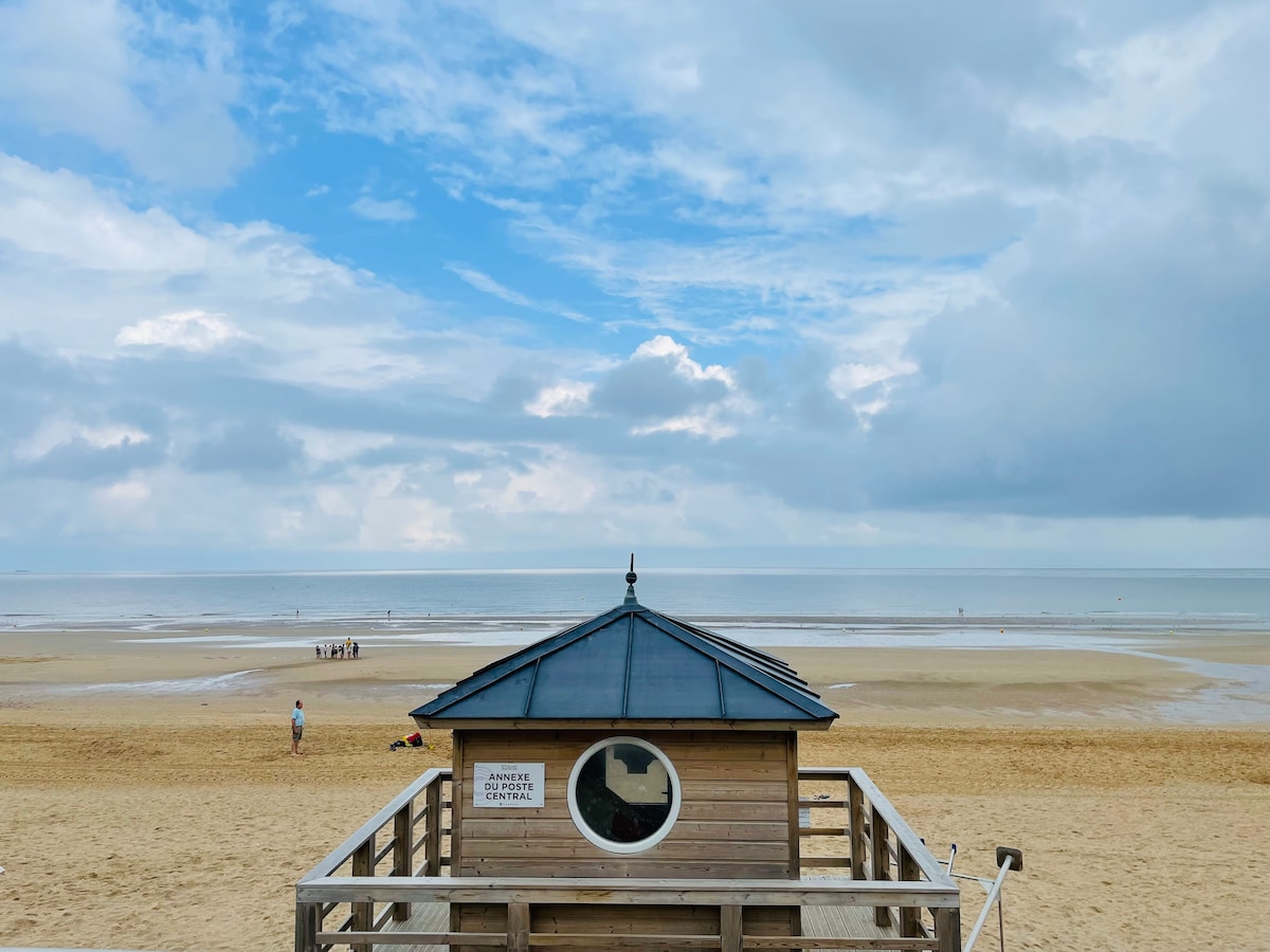 A wooden beach hut is positioned on a sandy shoreline, offering views of the calm sea and cloudy sky. Visitors can be seen enjoying the beach in the distance. The scene captures a tranquil coastal atmosphere, inviting relaxation and reflection.
