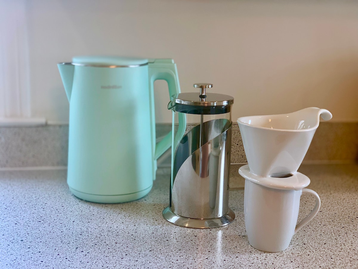 The image displays a modern mint green electric kettle alongside a stainless steel French press and a white ceramic coffee filter sitting atop a white mug. The items are arranged neatly on a light-colored countertop.