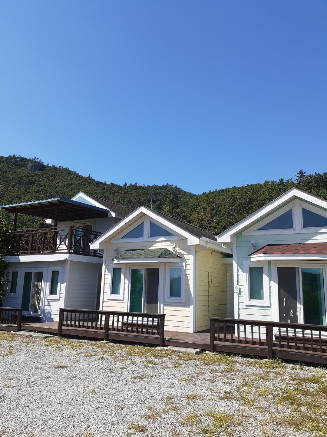 A charming cabin exterior is shown, featuring gable roofs and large windows. The building is surrounded by a gravel area and flanked by lush green hills in the background. A covered balcony is visible above, providing additional outdoor space.