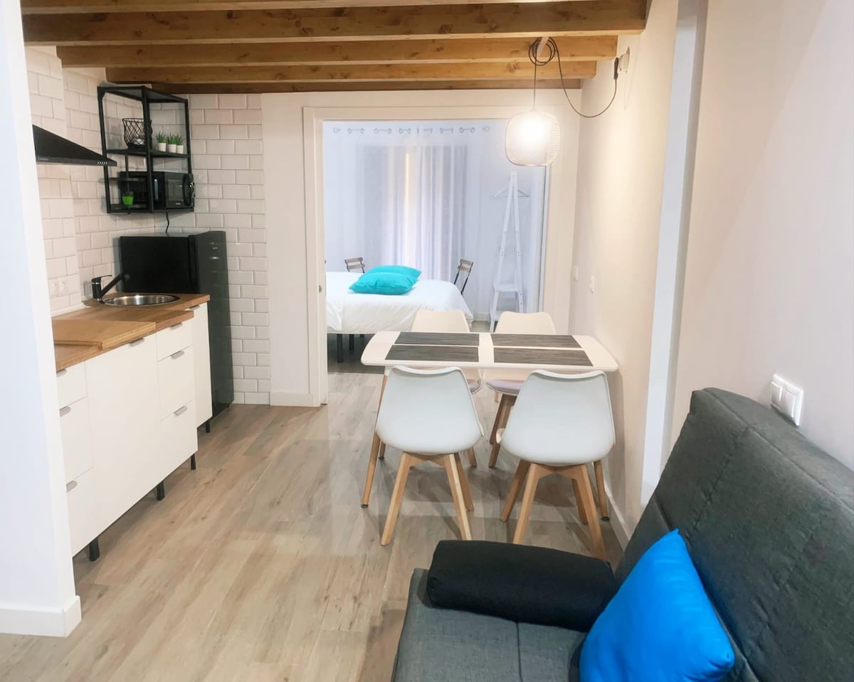 An open living space features a modern kitchen with a small sink and stovetop. A dining area is highlighted by a white table and chairs, while a gray sofa is complemented by a bright blue cushion. A partitioned sleeping area is visible in the background.