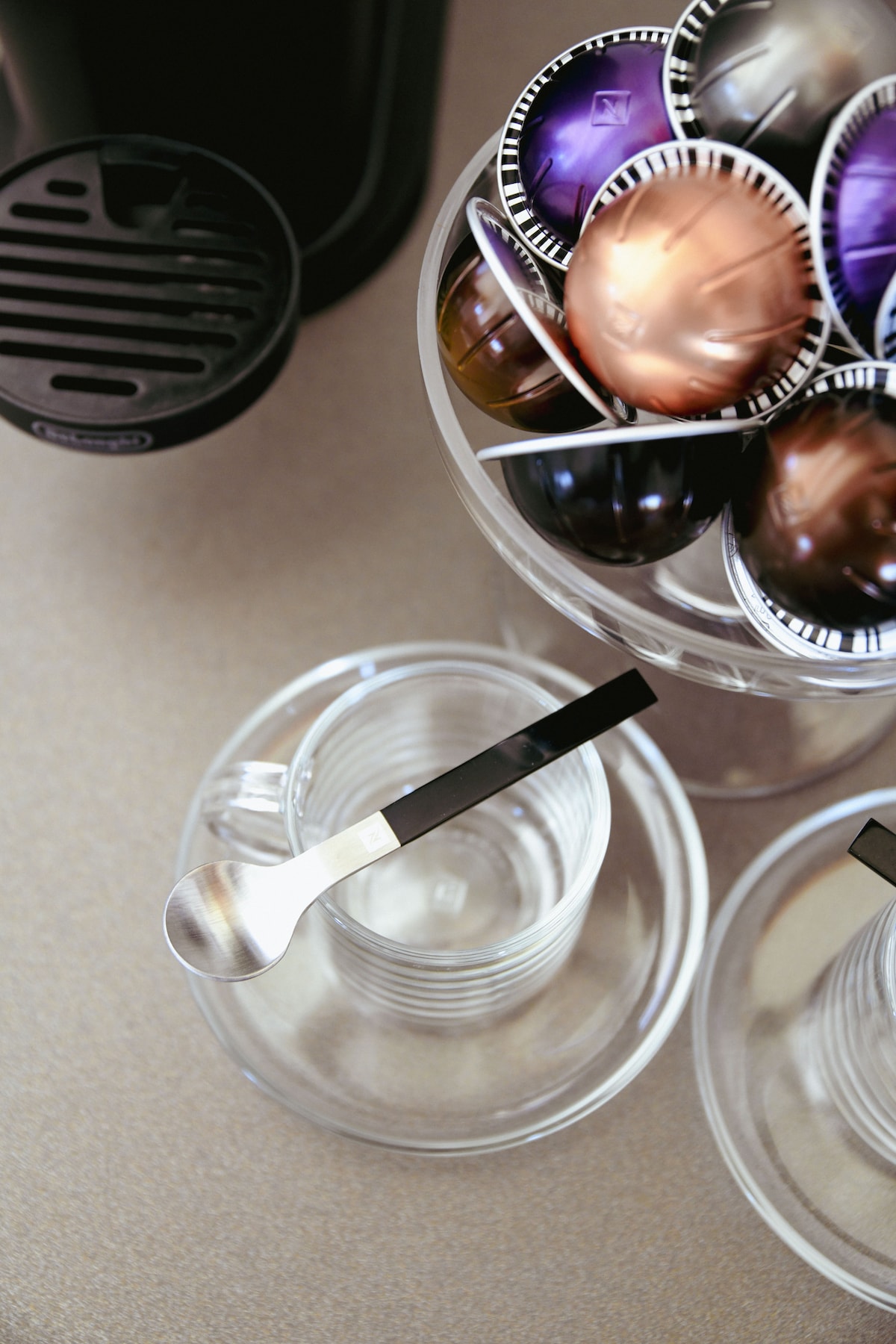 A modern coffee station is displayed, featuring an array of colorful coffee capsules organized in a clear bowl. A transparent cup sits nearby, accompanied by a sleek metallic spoon, highlighting the minimalist design of the setup.