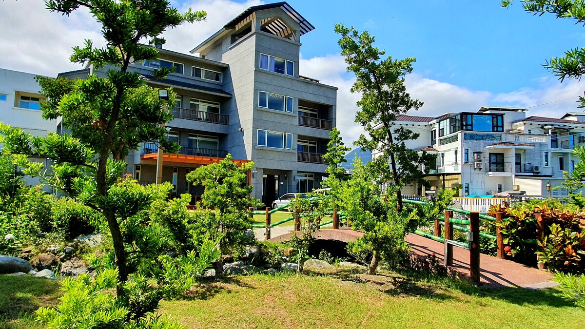 A modern building with multiple stories is set against a clear blue sky. Lush greenery surrounds the structure, with manicured shrubs and trees framing the scene. A footpath leads through the garden, inviting leisurely strolls and exploration of the serene outdoor space.