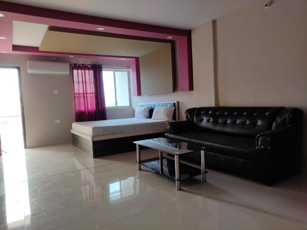 A spacious room is furnished with a comfortable seating area featuring a sleek black sofa and a glass coffee table. Natural light enters through a large window, complemented by burgundy curtains, while a cozy bed is set against a background of soft wall colors.