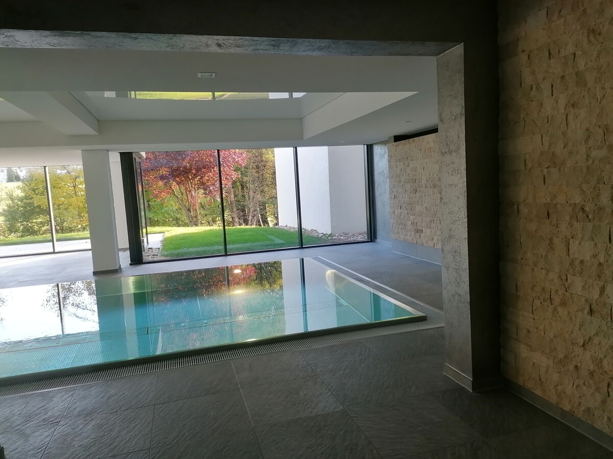 An indoor pool area is framed by large glass windows, allowing natural light to flood the space. The pool is filled with clear water, reflecting the greenery outside. Smooth stone walls enhance the modern ambiance, creating a serene environment for relaxation.