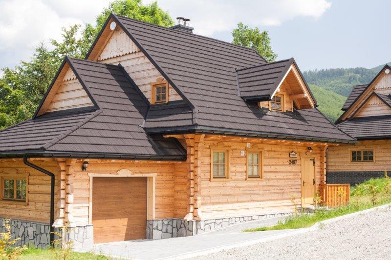 A charming wooden cabin showcases a traditional architectural style with a sloped roof and natural wooden exterior. A garage entrance is visible on the left side, complemented by a clear sky and green surroundings.