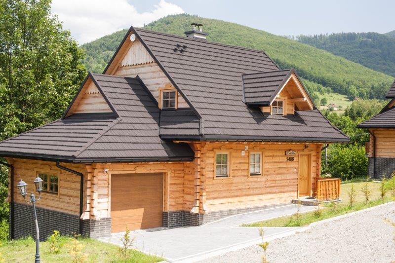 A charming wooden cabin is depicted with a sloped roof, featuring a combination of light wood and dark accents. The structure includes a garage and a welcoming front porch, set against a backdrop of lush green hills and clear blue skies.