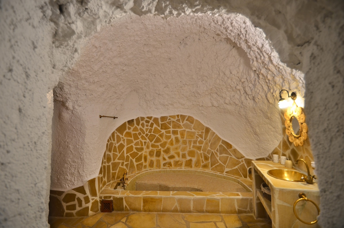 A bathroom with a unique cave-like design features a stone bathtub, complemented by a decorative stone wall. A sink with a vintage-style mirror is positioned next to the tub, creating a serene and distinctive bathing space.