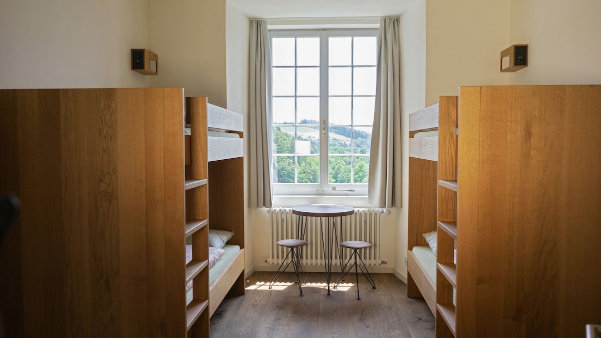 A bright room features two wooden bunk beds positioned against the walls, providing ample space. A small table with two chairs is situated in front of a large window, offering views of the greenery outside. Neutral tones and hardwood floors complete the serene environment.