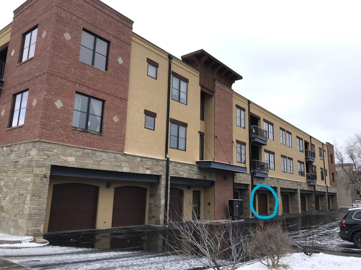 A modern condo building with a mix of brick and stucco exterior is shown, featuring stone accents at the base. Several balconies overlook the street, and a private garage is visible at the ground level, offering access to the living spaces above.