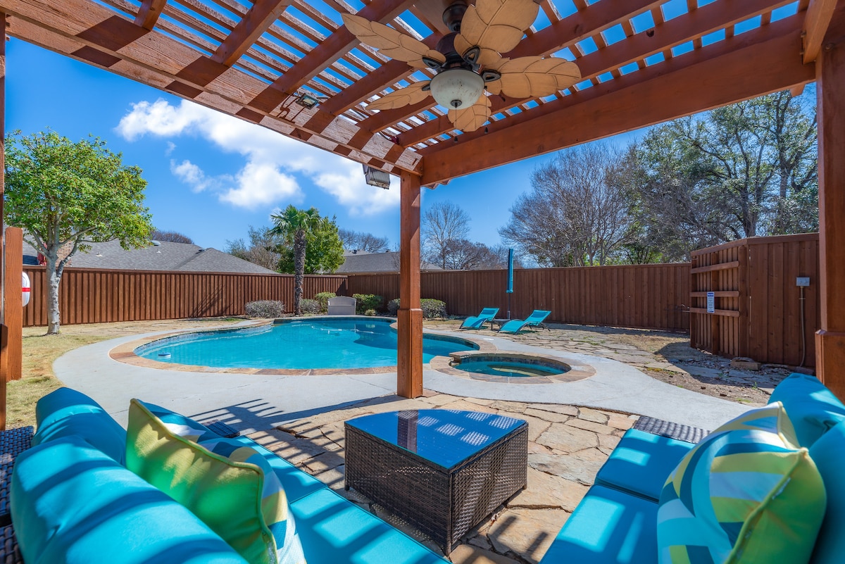 An outdoor pergola provides shaded seating, with plush cushions overlooking a sparkling pool and heated spa. Sun loungers are positioned for relaxation, and surrounding trees offer a touch of greenery, complementing the spacious patio area.