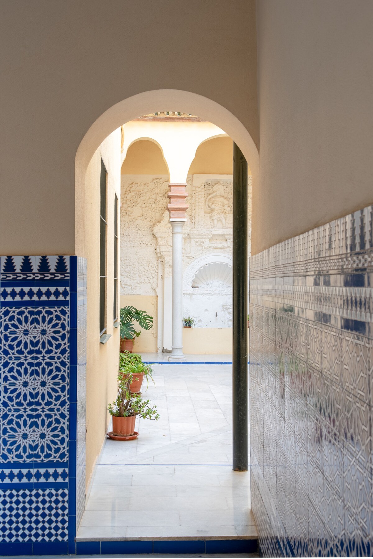 A soft, natural light filters through an arched passageway, revealing a serene courtyard adorned with ornamental tiles. Lush plants are positioned in terracotta pots against textured walls, adding greenery to the inviting space, which features a decorative wall with carved details.